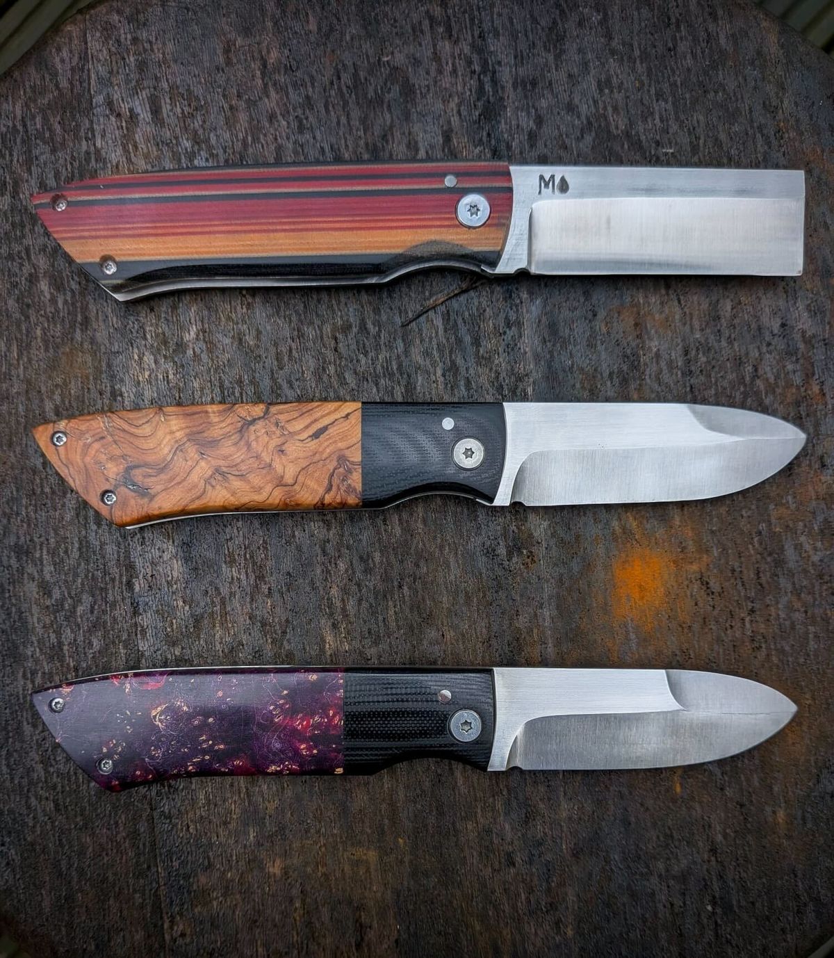 Three handmade folding knives with blades open, featuring striped wood, olive wood, and resin handles